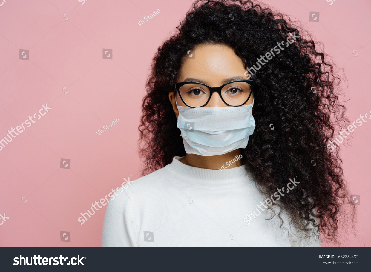 Covid-19, infectious virus. Close up shot of young woman with curly bushy hair, wears transparent glasses and medical disposable mask, cares about her health, protects in dangerious situation #1682884492