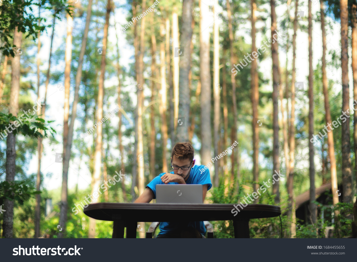 A man working remotely on his laptop on a table outdoor among the trees. It looks like he works in a jungle #1684455655