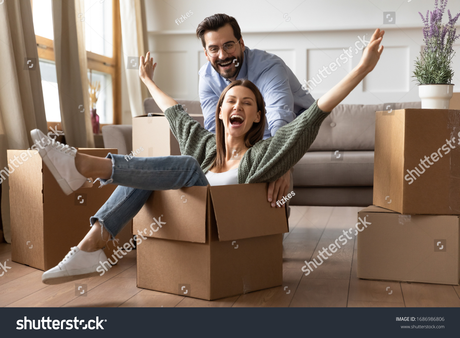 Full length overjoyed young bearded man in glasses pushing laughing wife in carton box. Energetic happy woman sitting in carboard container, having fun with smiling husband in new apartment house. #1686986806