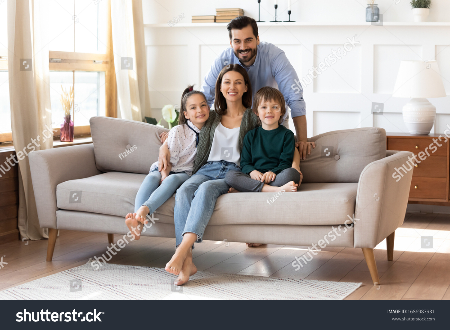 Portrait of smiling bearded father standing near couch with sitting happy wife and little children. Joyful affectionate family of four posing for photo, looking at camera, good relations concept. #1686987931