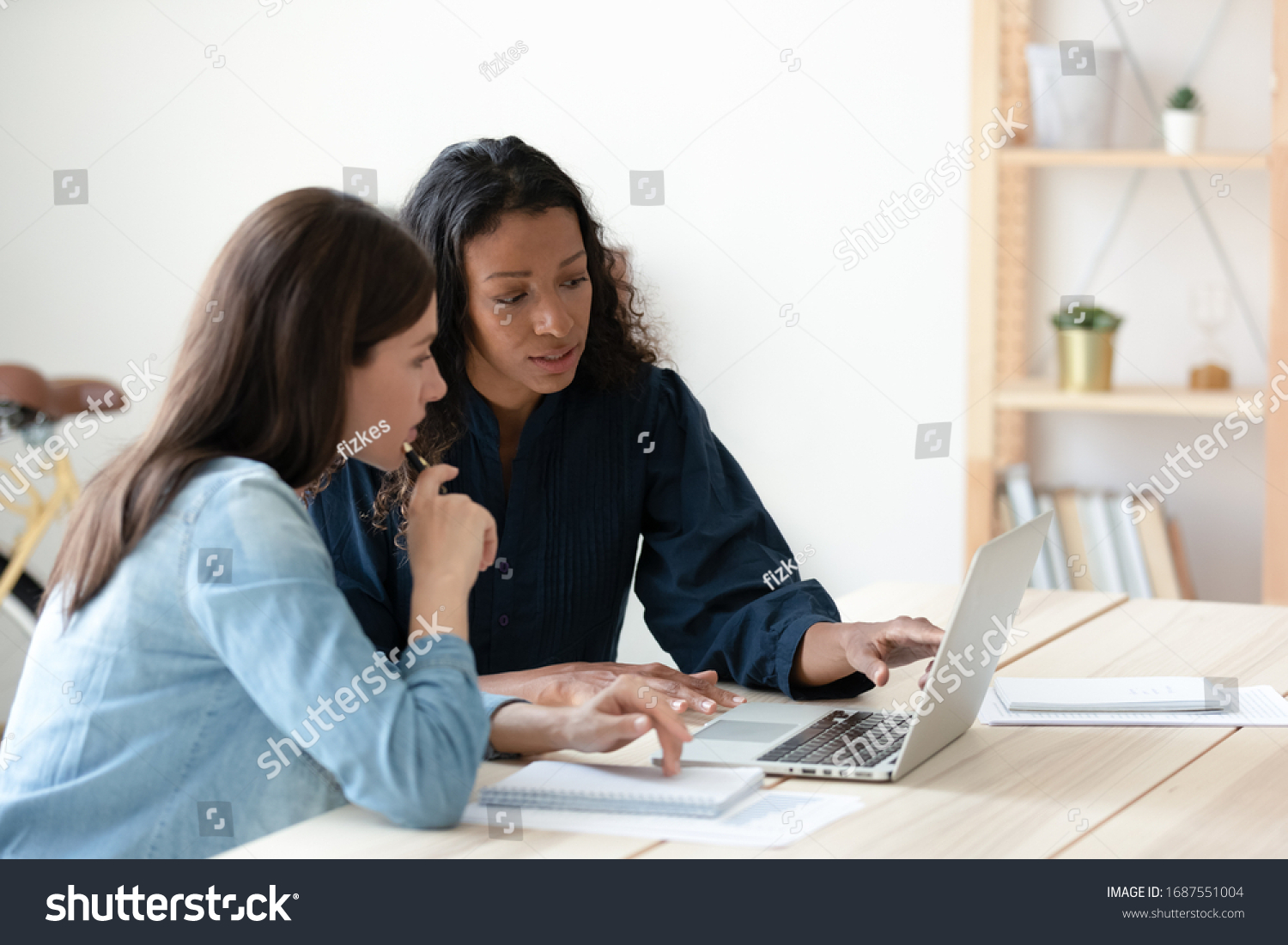 Focused diverse female colleagues sit at desk in office brainstorm discuss business idea use laptop, concentrated woman coworkers look at computer screen work together at meeting, cooperation concept #1687551004