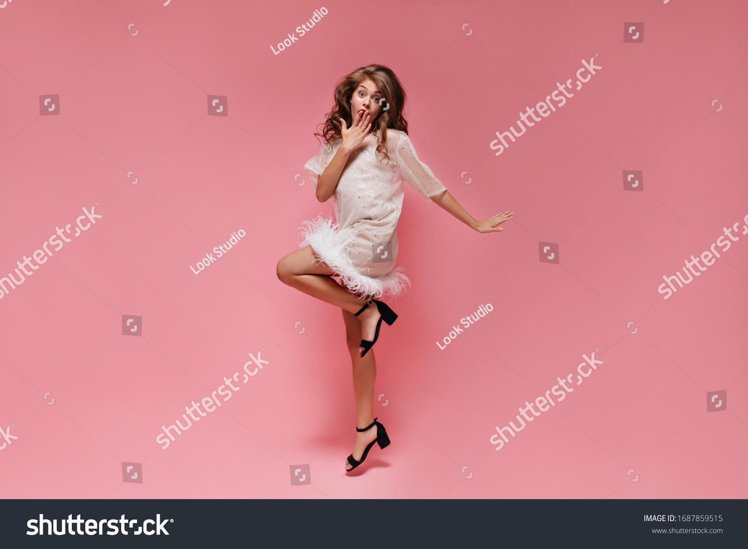 Surprised woman in white dress jumps on pink background. Happy shocked girl covers mouth and moves on isolated. #1687859515