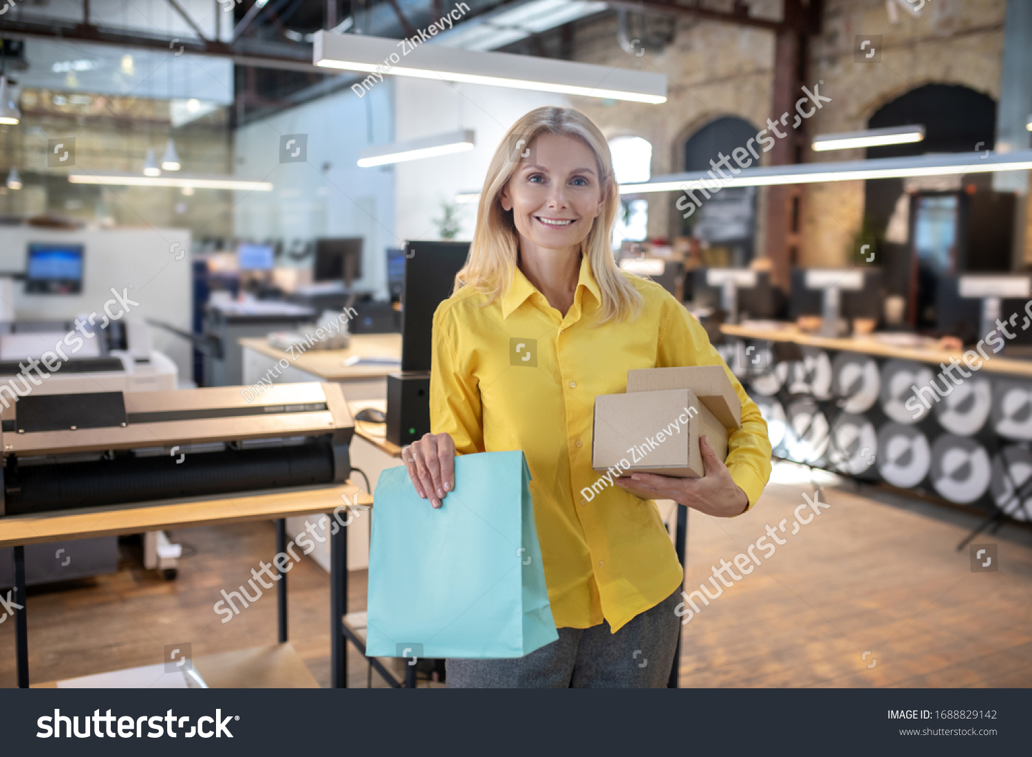 At workshop. Blonde woman holding boxes and paper packege, smiling #1688829142