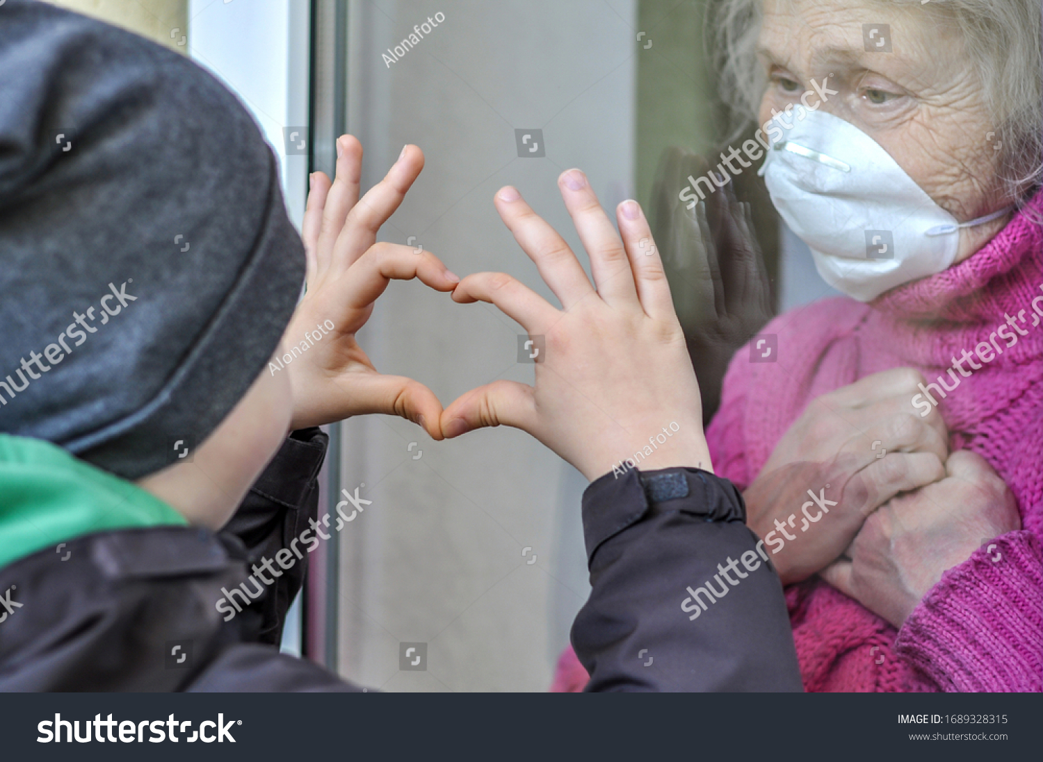 Grandmother mature woman in a respiratory mask communicates with her grandchild through a window. Elderly quarantined, isolated. Coronavirus covid-19. Caring with older people. Family values, love #1689328315