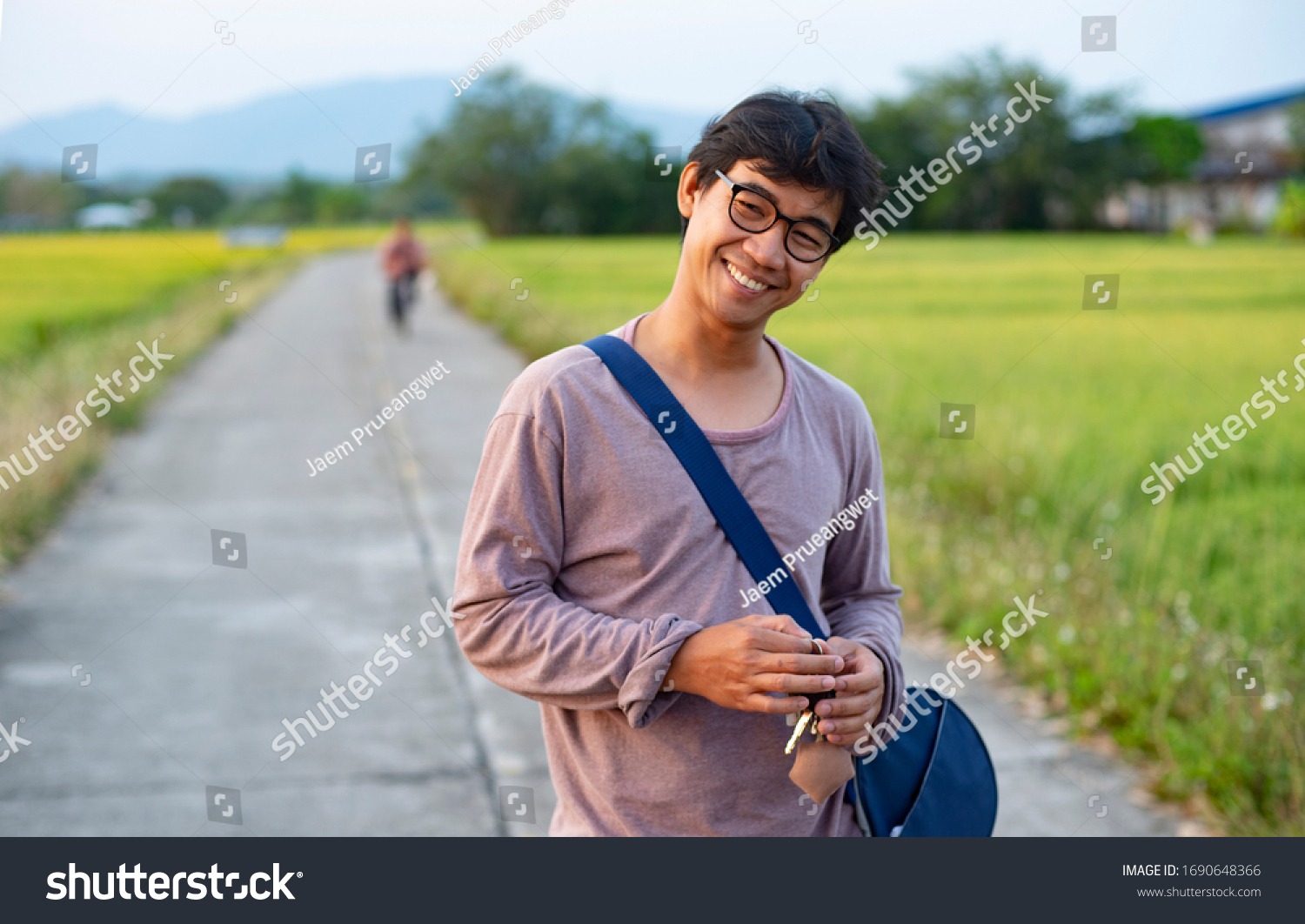 Portrait of an Asian Thai man looking humble and relaxed surrounded by rice field and in the background there is an unknown person riding a bicycle #1690648366