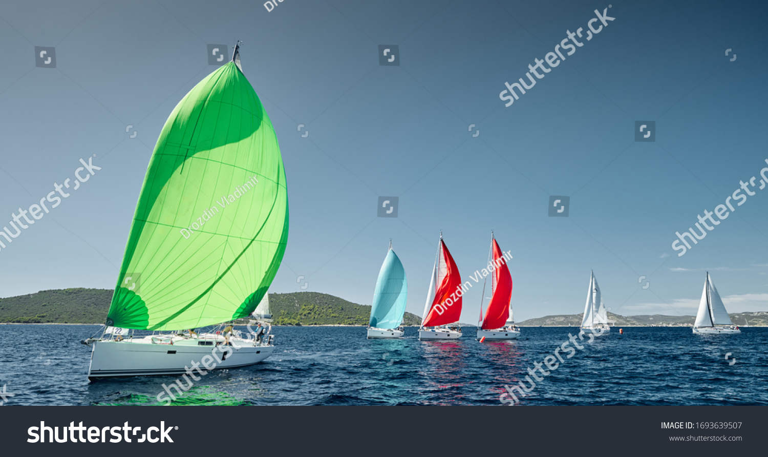 Sailboats compete in a sail regatta at sunset, race of sailboats, reflection of sails on water, multicolored spinnakers, number of boat is on aft boats, island is on background, clear weather #1693639507