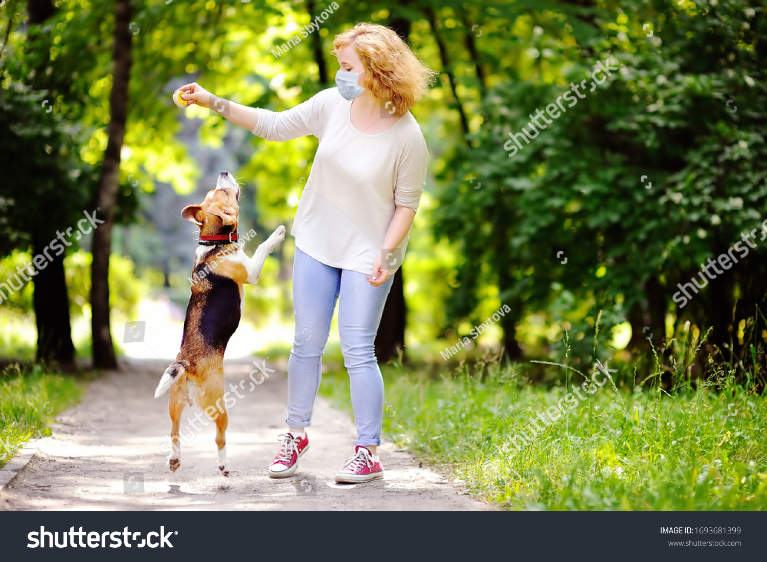 Young beautiful woman wearing disposable medical face mask playing with Beagle dog in the park during coronavirus outbreak. Walking of pets. Safety in a public place while epidemic of covid-19. #1693681399