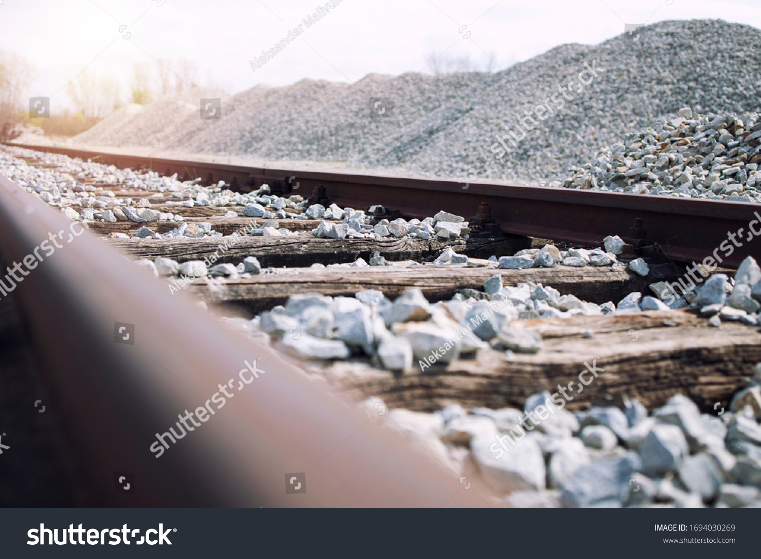 Railroad construction site. Railway tracks with wooden rail ties and ballast. In background large pile of ballast rocks used for building railroads. #1694030269