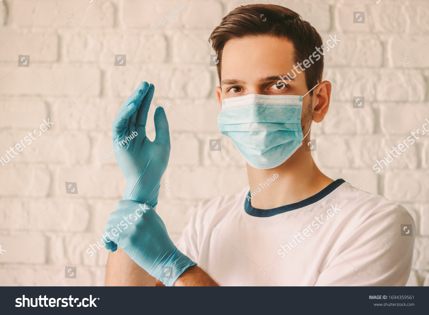 Confident man surgeon in medical mask on face wearing protective sterile glove on hand. Professional doctor in protective face mask putting on latex glove on hand. Coronavirus COVID-19. Hospital staff #1694359561