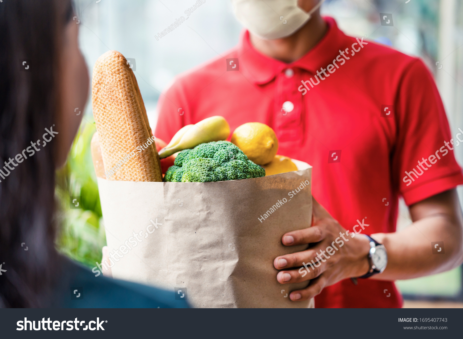 Asian deliver man wearing face mask in red uniform handling bag of food, fruit, vegetable give to female costumer in front of the house. Postman and express grocery delivery service during covid19. #1695407743