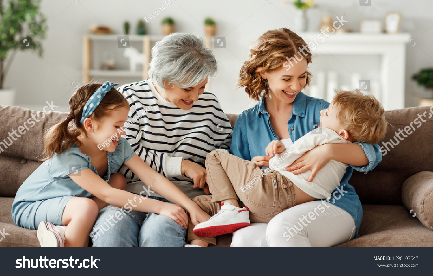 Grandmother and mother hugging and tickling laughing boy while sitting on couch and playing with children on weekend day at home
 #1696107547