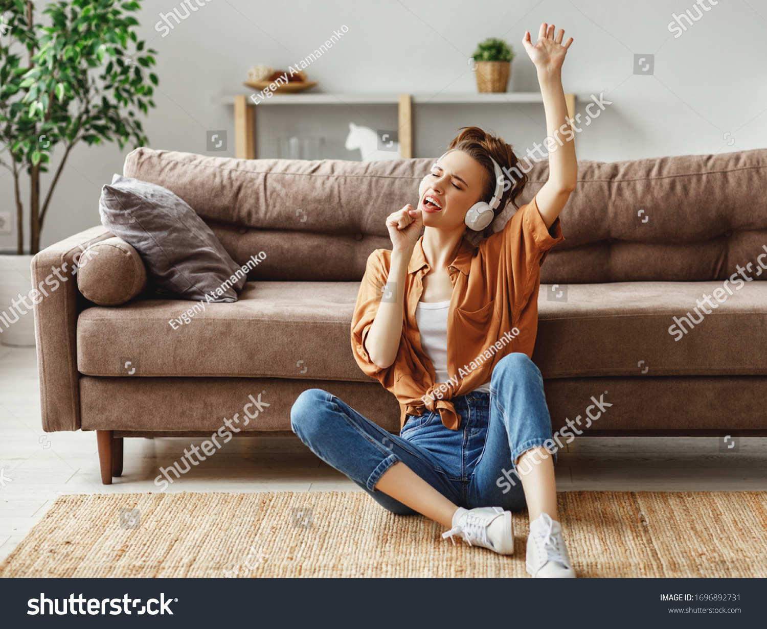 Young female in casual outfit listening to music in headphones and singing while sitting on floor near sofa and having fun at home
 #1696892731