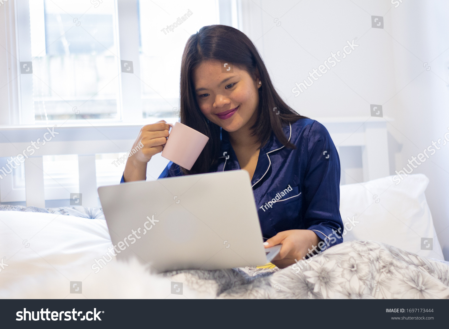 Young asian smiling woman working on laptop while holding a pink mug. Happy morning #1697173444