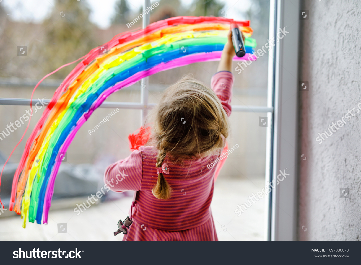 Adoralbe little toddler girl with rainbow painted with colorful window color during pandemic coronavirus quarantine. Child painting rainbows around the world with the words Let's all be well. #1697330878