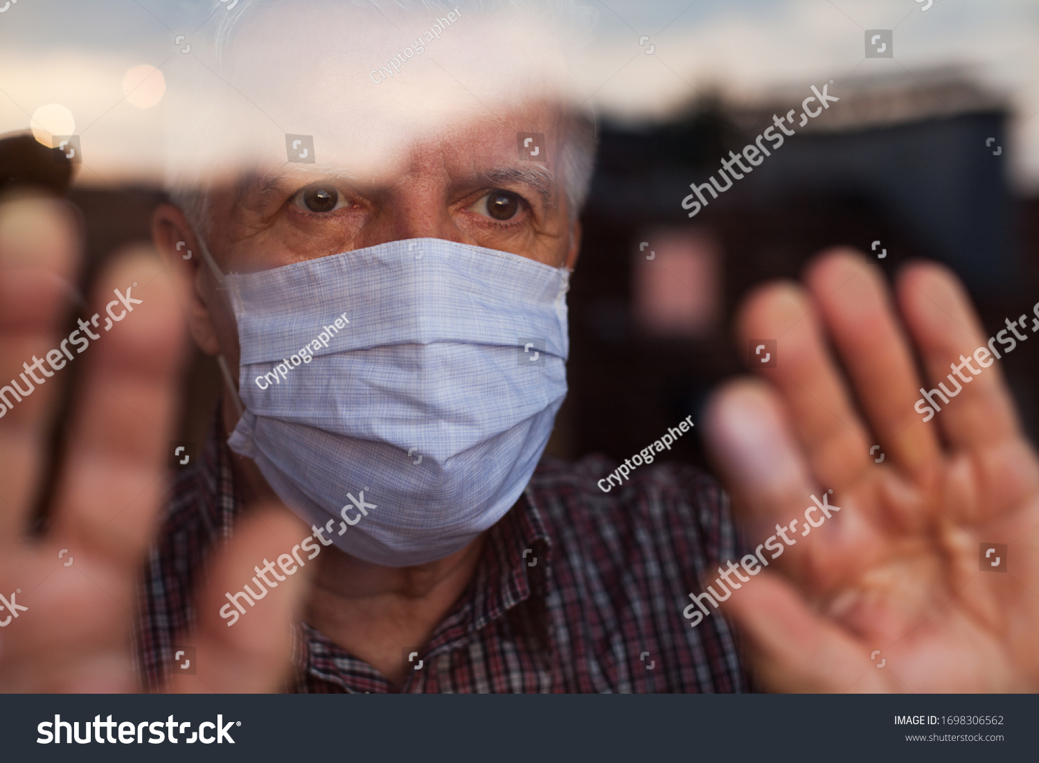 Elderly caucasian man wearing hand made protective face mask,in nursing care home,looking outside window, sadness,stress & hope in his eyes,self isolation due to global COVID-19 Coronavirus pandemic