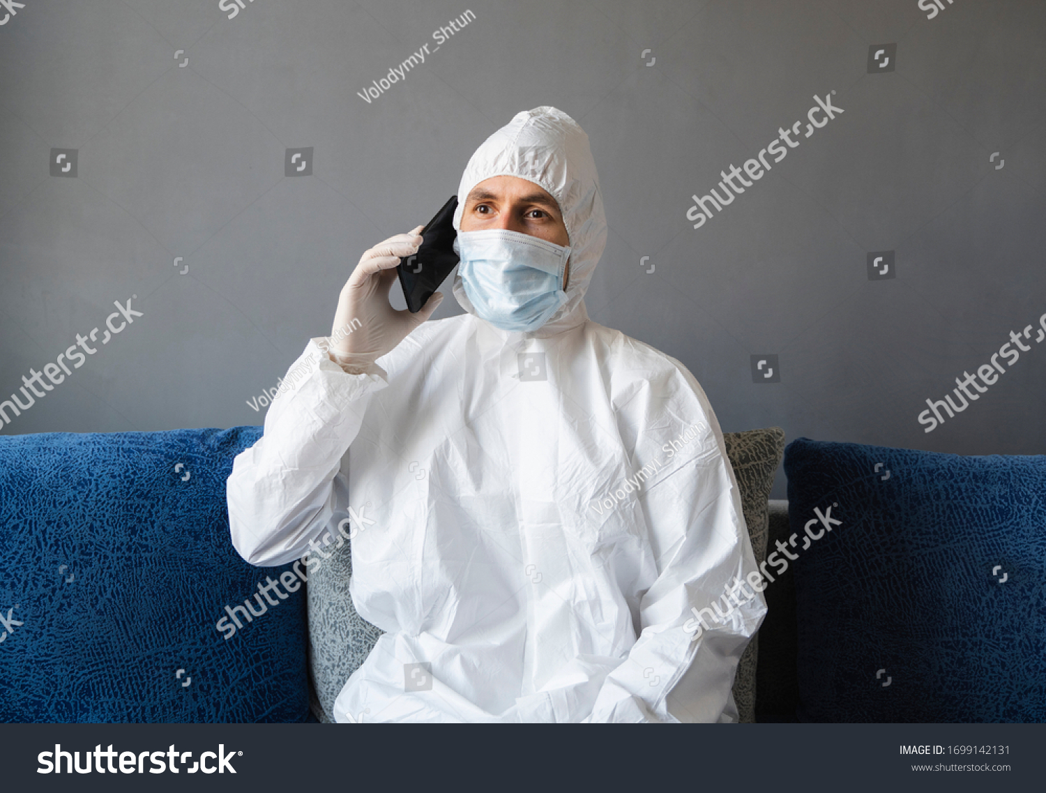 Man in protective suit, medical mask and rubber gloves sits at home and speaking with a friends or business partners on a sofa during quarantine. Man at remote work in a pandemic covid. #1699142131