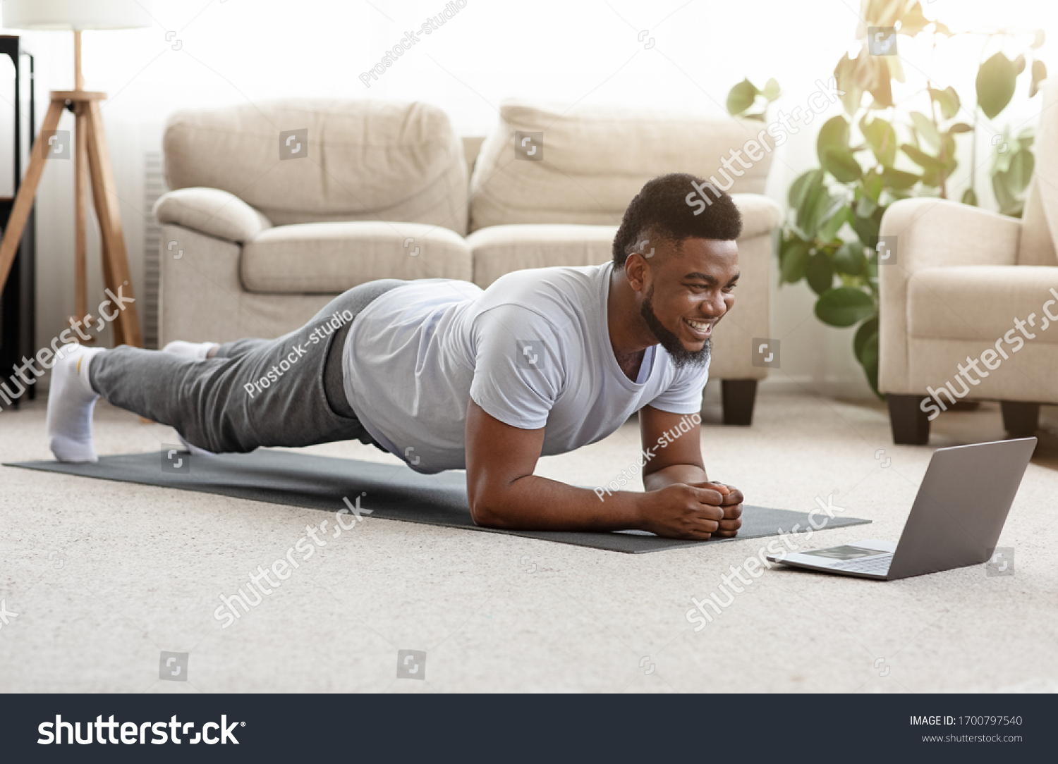 Training At Home. Sporty man doing yoga plank while watching online tutorial on laptop, exercising in living room, free space #1700797540
