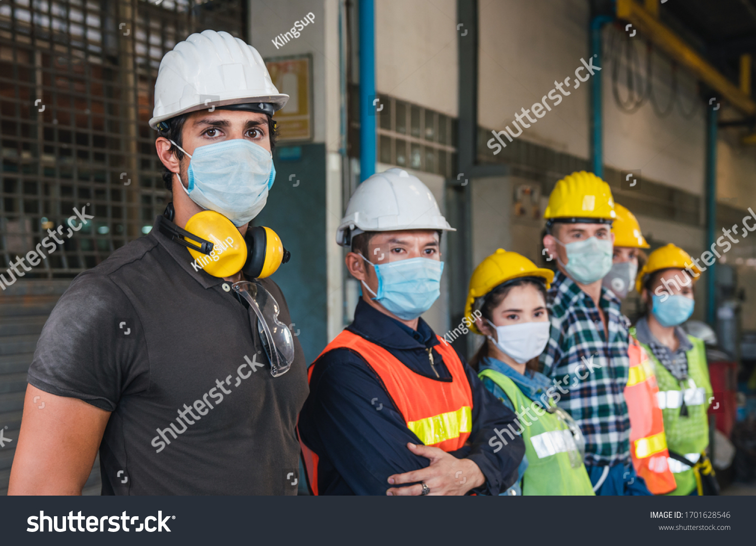 Group of diverse team of workers wearing face mask and protective helmets standing in front of the factory #1701628546