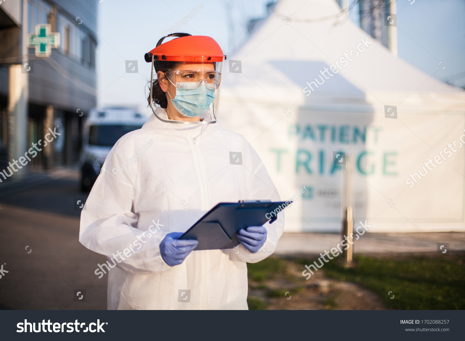 Paramedic wearing personal protective equipment PPE holding folder standing in front of ICU hospital isolation rt-PCR drive thru testing site,COVID-19 pandemic outbreak crisis,worried exhausted staff #1702088257