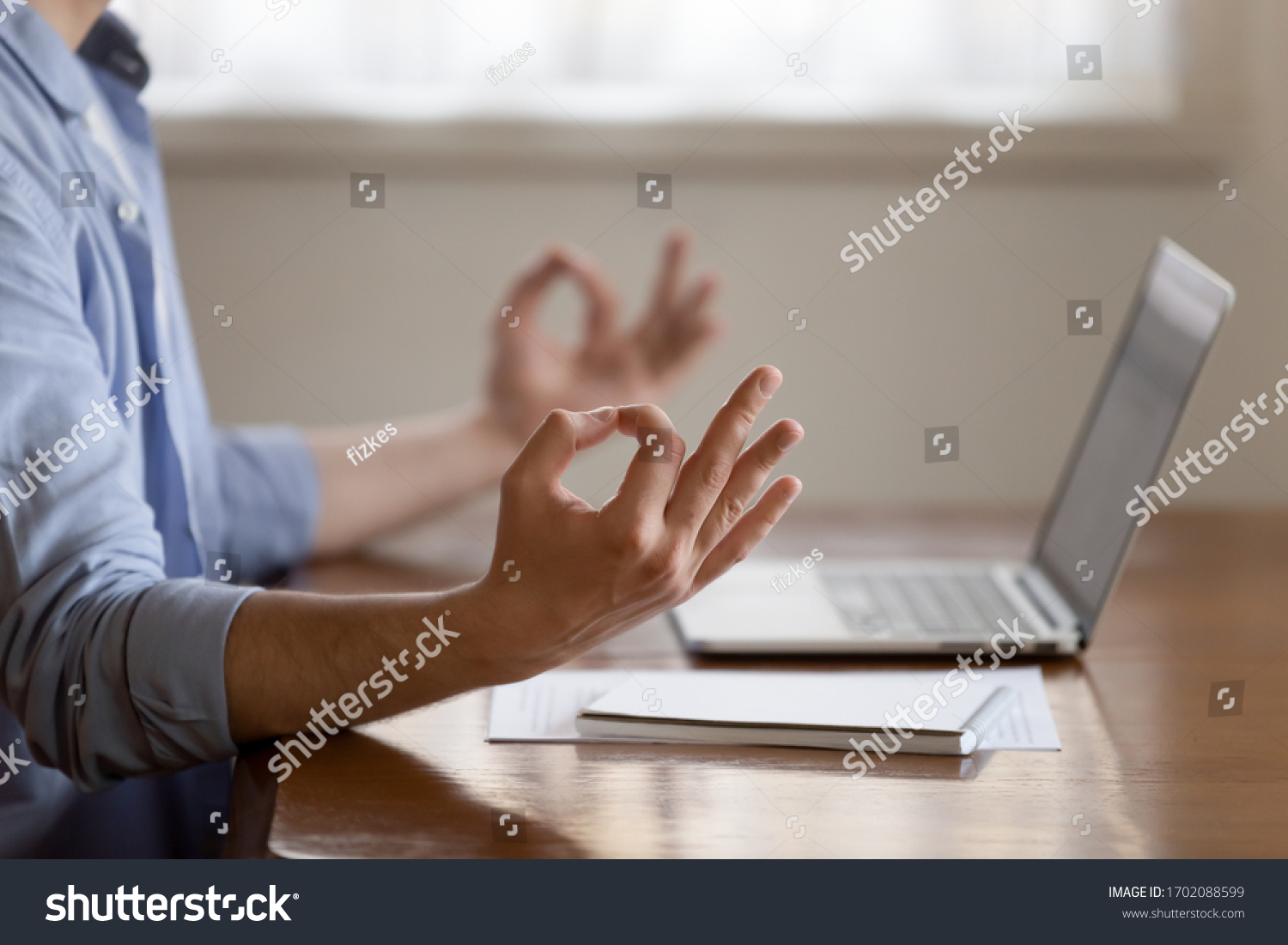 Close up focus on folded in mudra gesture male hands. Young peaceful man managing stress at workplace, meditating, calming down, doing yoga breathing exercises, sitting at table alone indoors. #1702088599