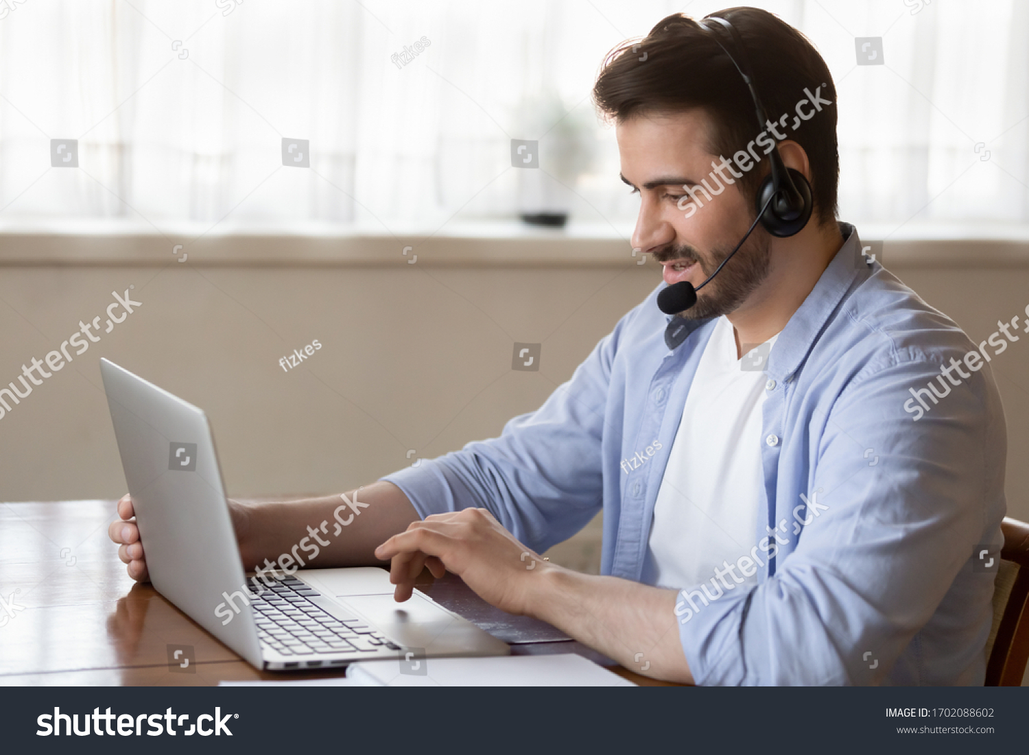 Side view young man wearing wireless headset with microphone, looking at laptop screen, study on online courses. Skilled male teacher giving online educational class to client, working remotely. #1702088602