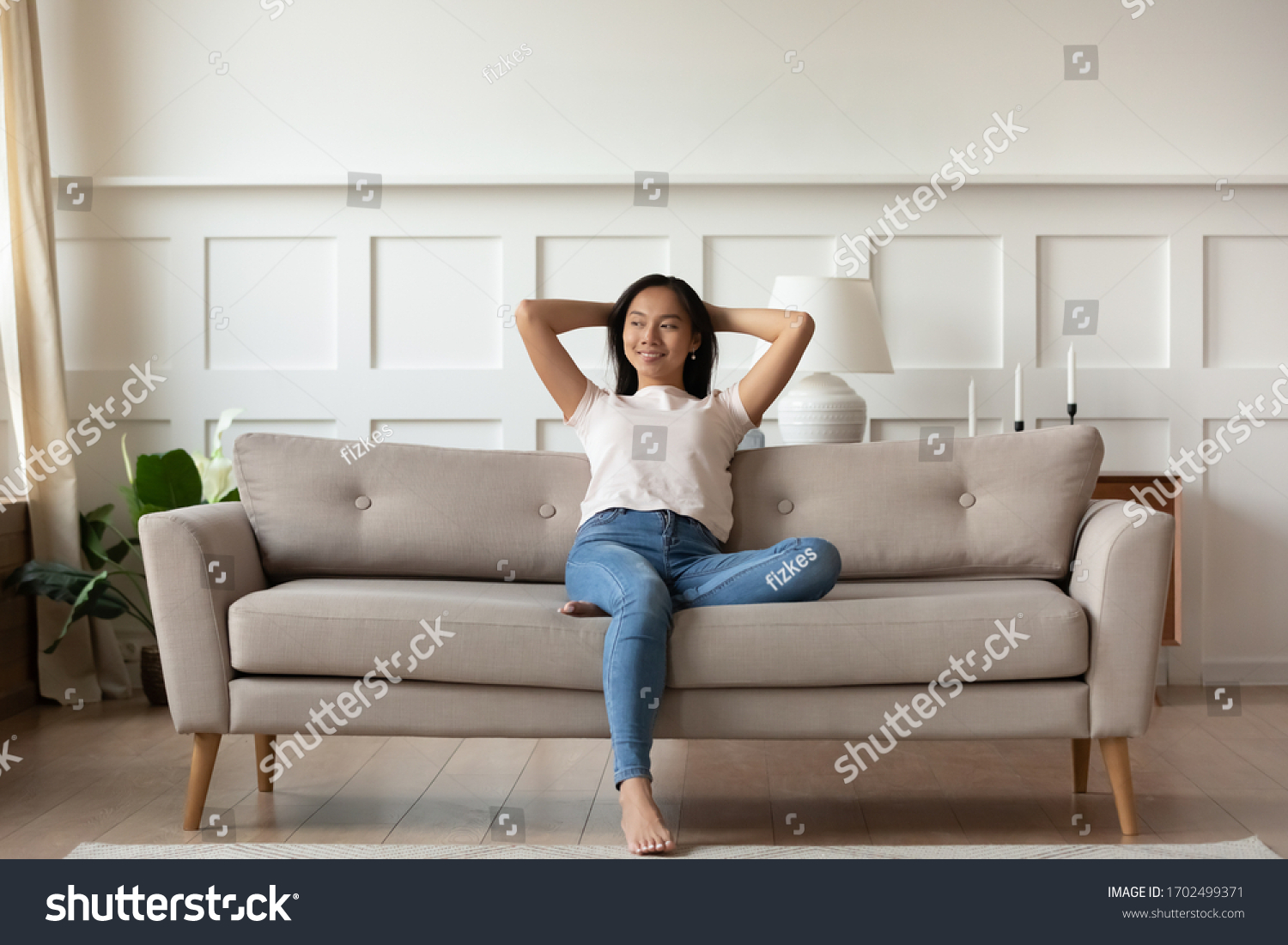 Asian woman looks at distance resting leaned on couch enjoy fresh air in summer day in modern fashionable living room interior, full length image. Contemporary apartments owner or carefree day concept #1702499371