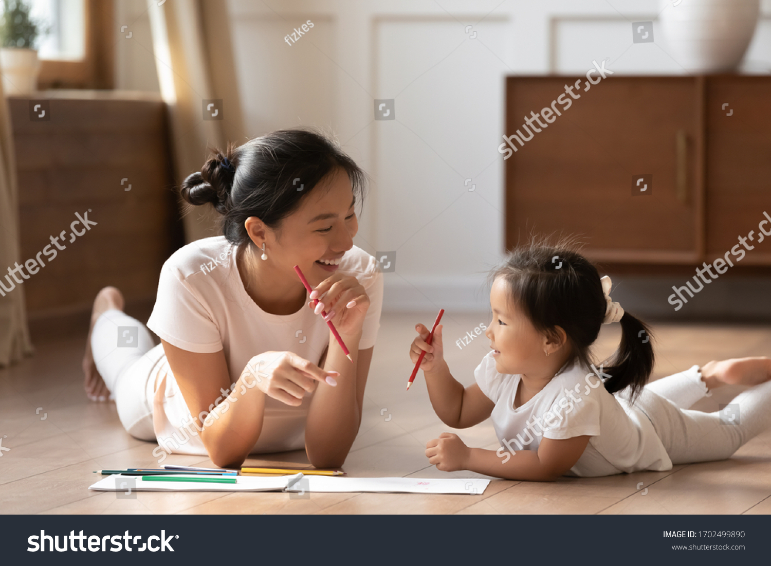 Educational pastime develop creativity skill in kid concept. Asian mother her small daughter lying on warm wooden floor in sunny cozy living room, mom teach girl paint use album and colourful pencils #1702499890