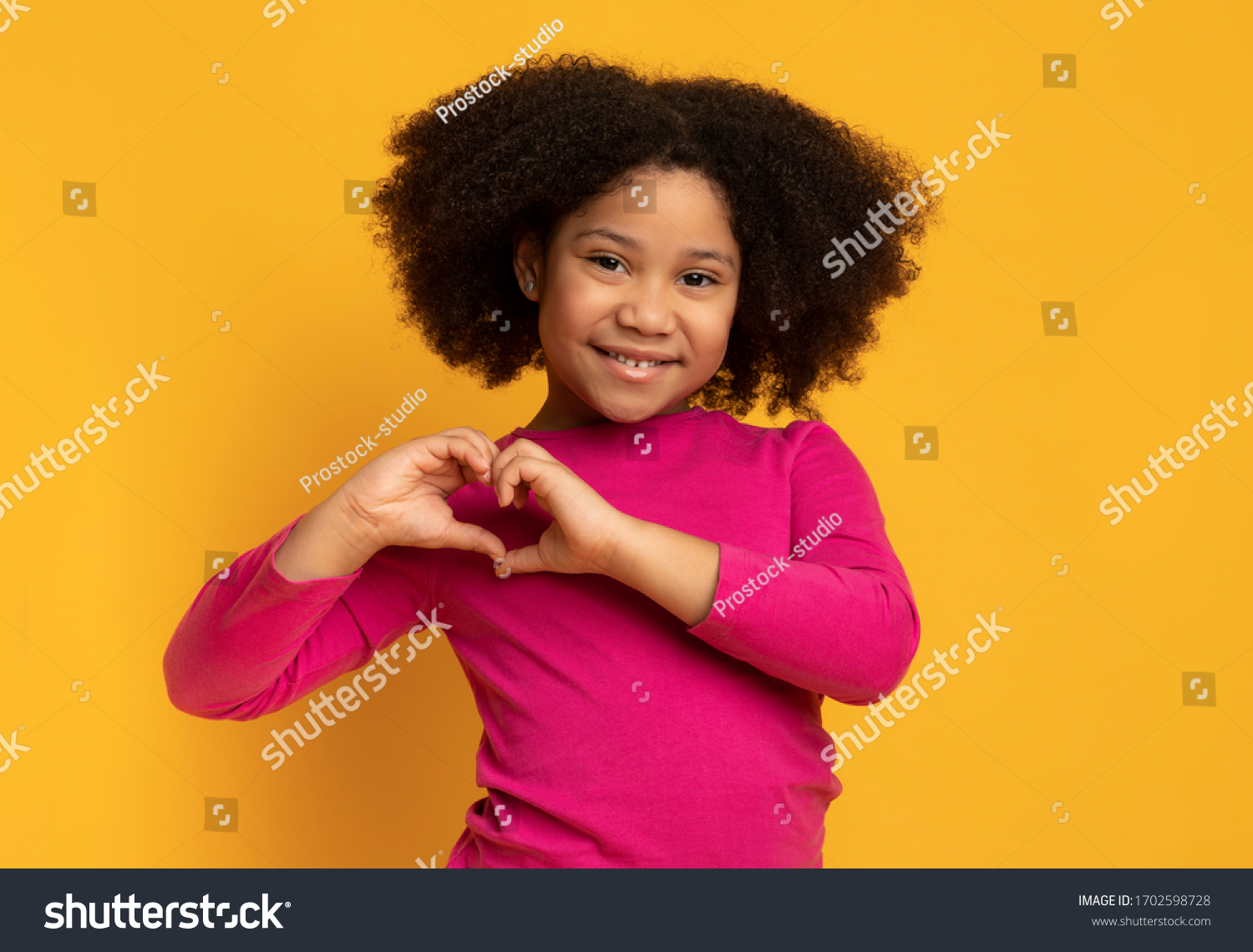 Cute little african american girl making heart shape with her hands and smiling to camera, posing over yellow background in studio, empty space #1702598728