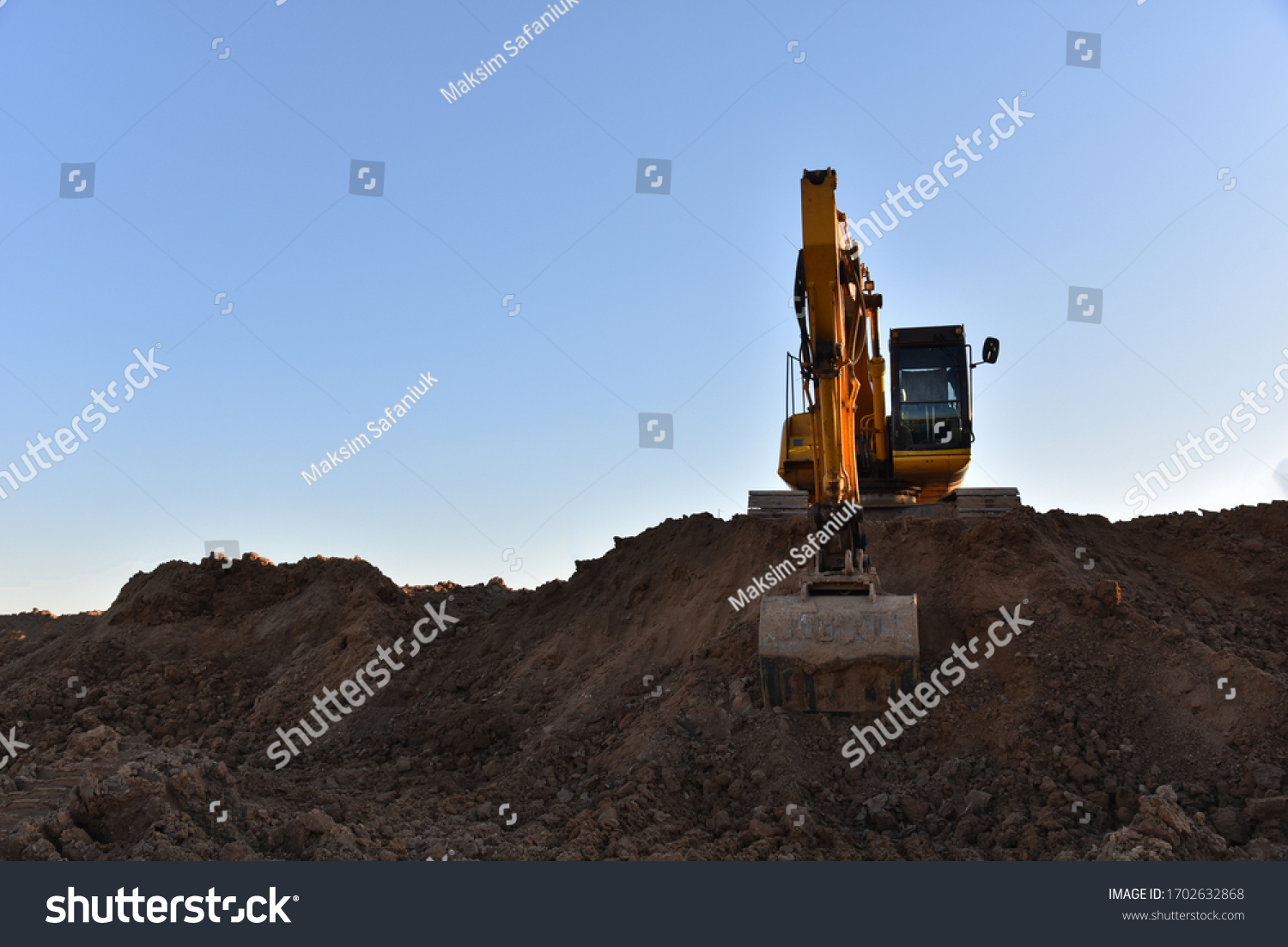 Yellow excavator during earthmoving at open pit on blue sky background. Construction machinery and earth-moving heavy equipment for excavation, loading, lifting and hauling of cargo on job sites #1702632868