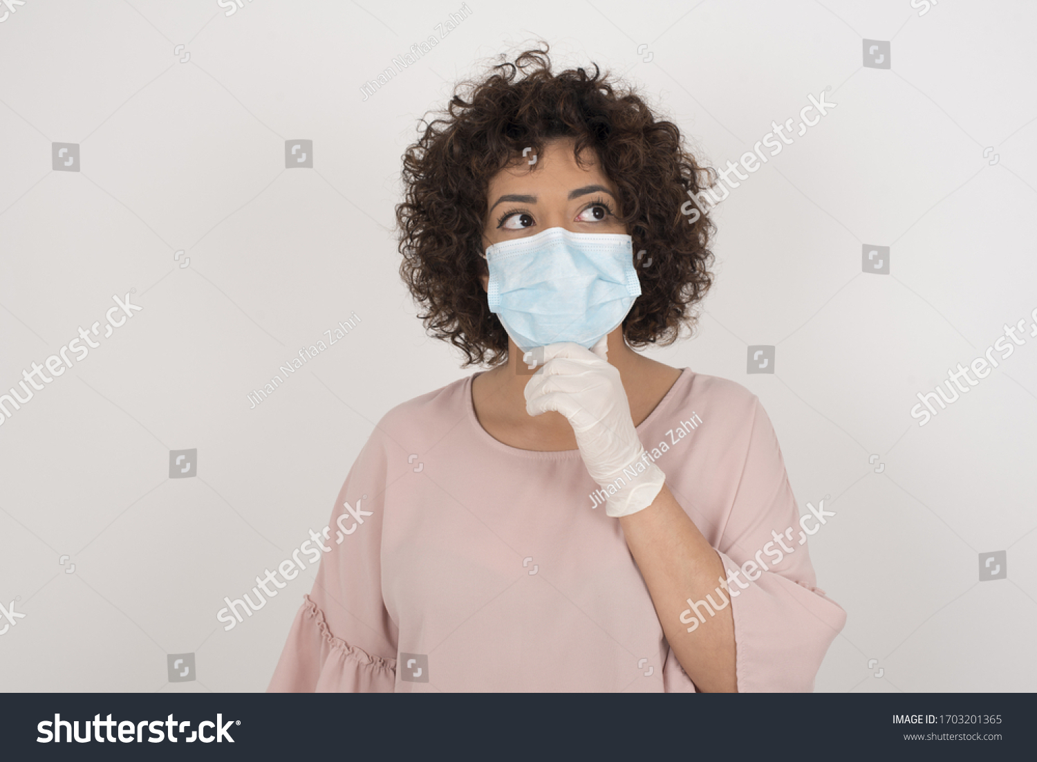 Shot of contemplative thoughtful Young woman keeps hand under chin, looks thoughtfully upwards, dressed in casual clothes. wearing face mask protection against infectious disease. #1703201365