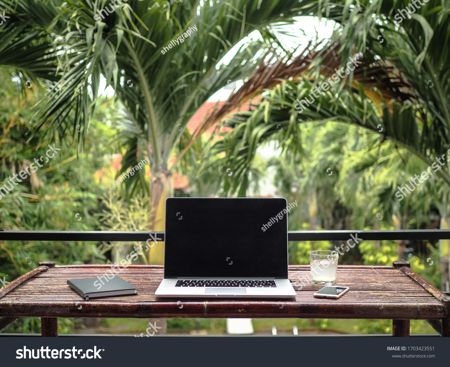 laptop of a remote digital nomad on a wooden bamboo table with notebook, mobile phone and glass in nature with a green tropical background with palm trees #1703423551