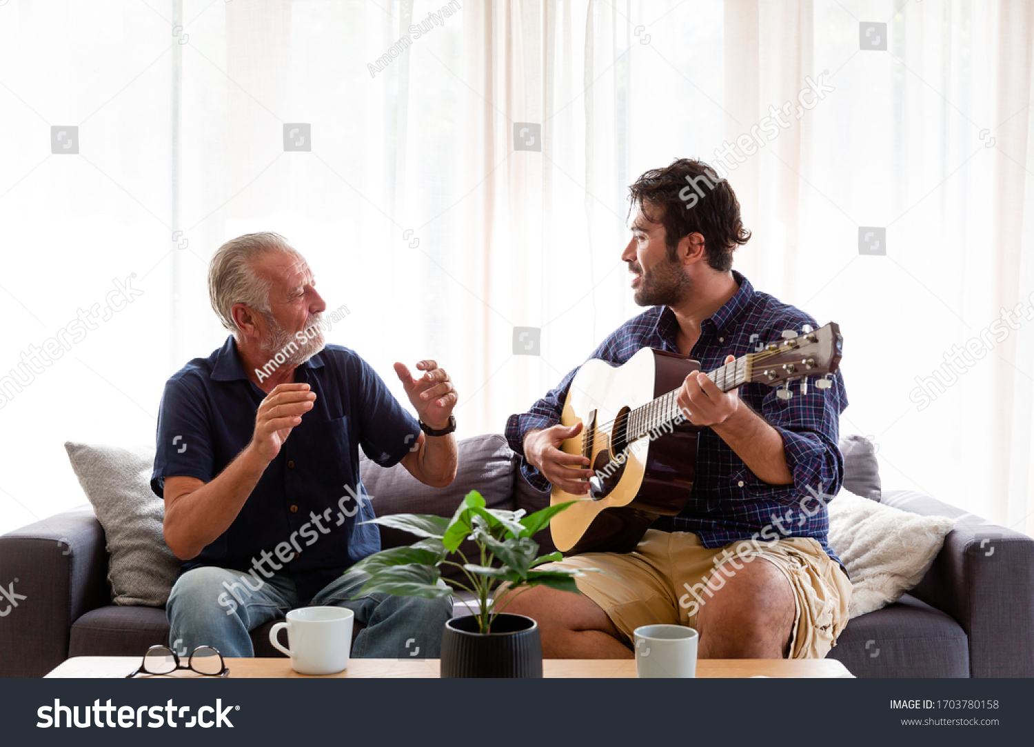 Happy Senior Father sing a song and Adult Son playing guitar on the sofa at home. Elderly father and son spending time together at home. #1703780158