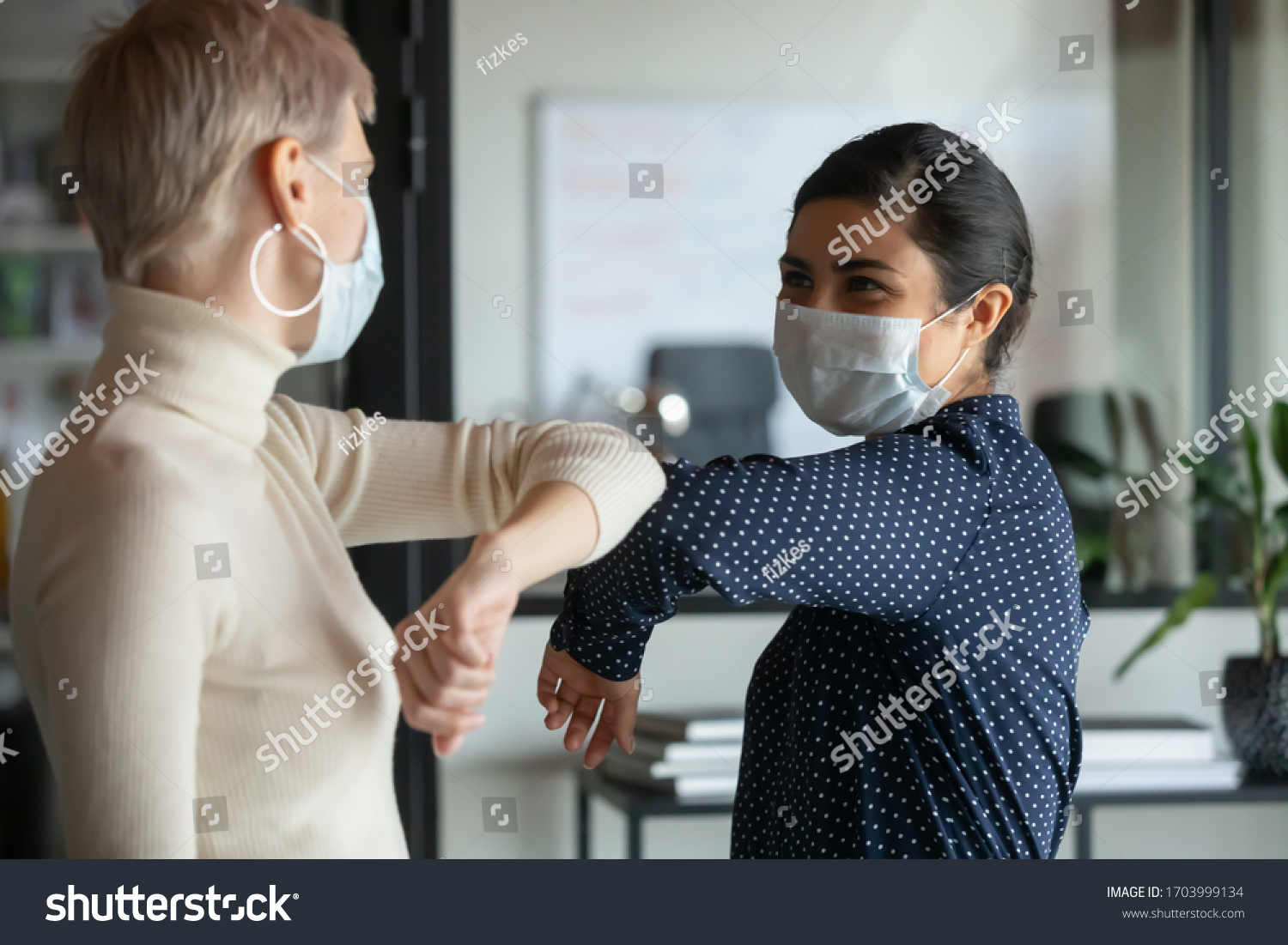 Smiling diverse female colleagues wearing protective face masks greeting bumping elbows at workplace, woman coworkers in facial covers protect from COVID-19 coronavirus in office, healthcare concept #1703999134
