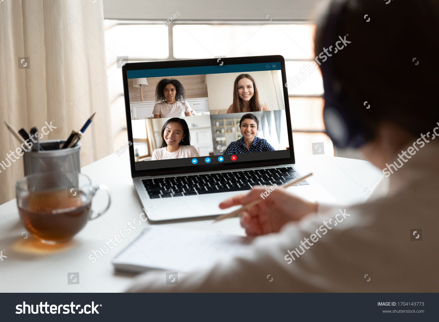 Woman sitting at desk noting writing information studying at home with multiracial students diverse ladies makes video call using video conference application, view over girl shoulder to laptop screen #1704143773