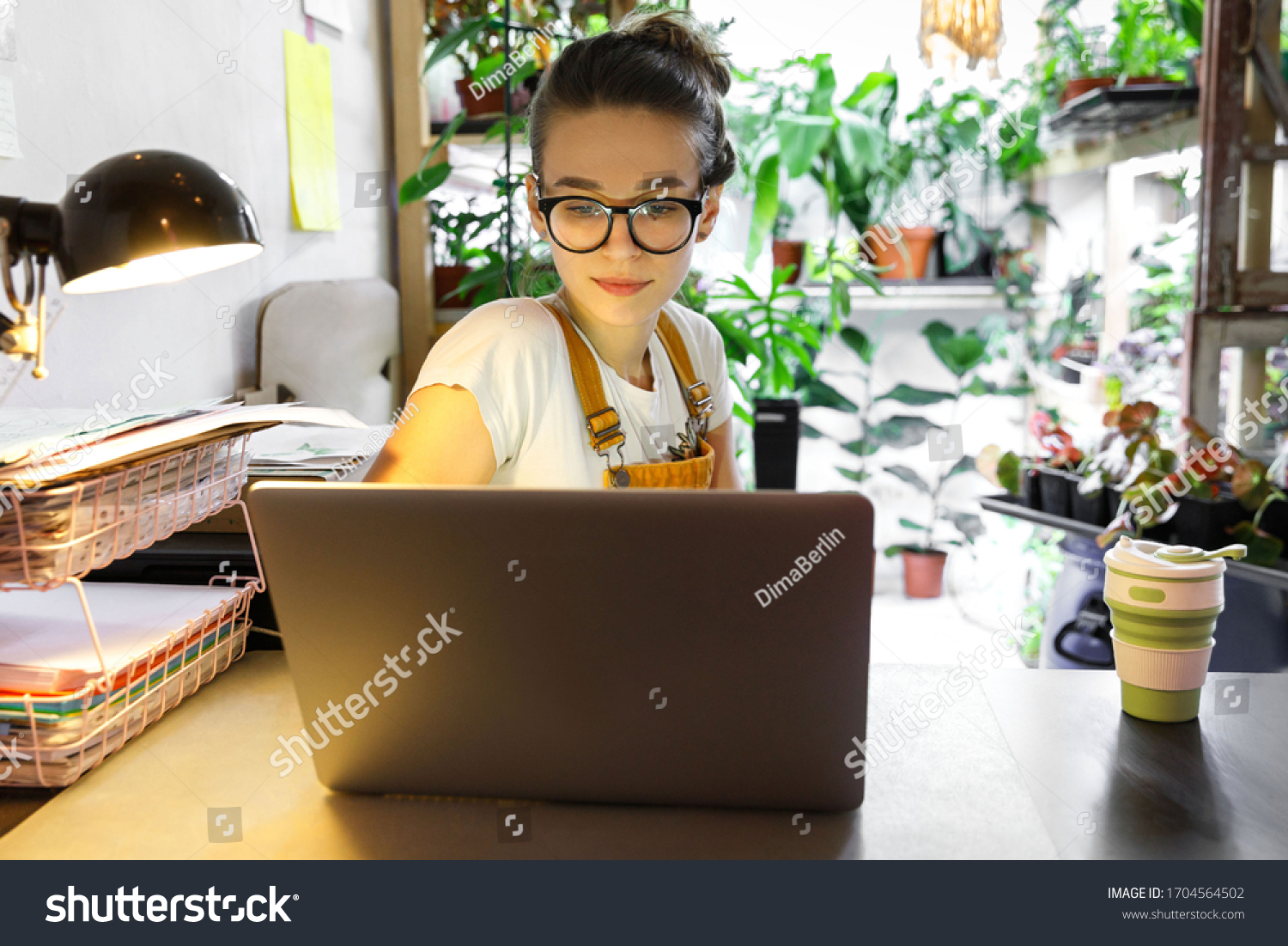 European female gardener in glasses using laptop, scrolling through social networks, reads news, coffee/tea mug on table, home garden/greenhouse on background. Cozy workplace, remote work #1704564502