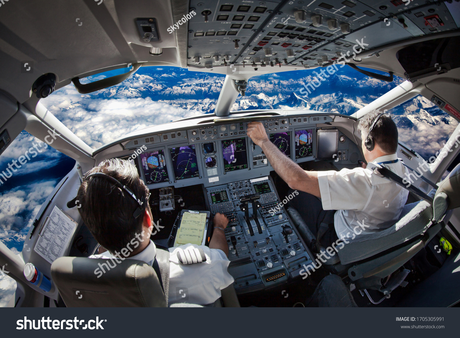 Cockpit of the modern passenger aircraft in flight. Pilots fly an airplane over the mountain landscape. Blue cloudy sky is visible outside the cockpit. #1705305991