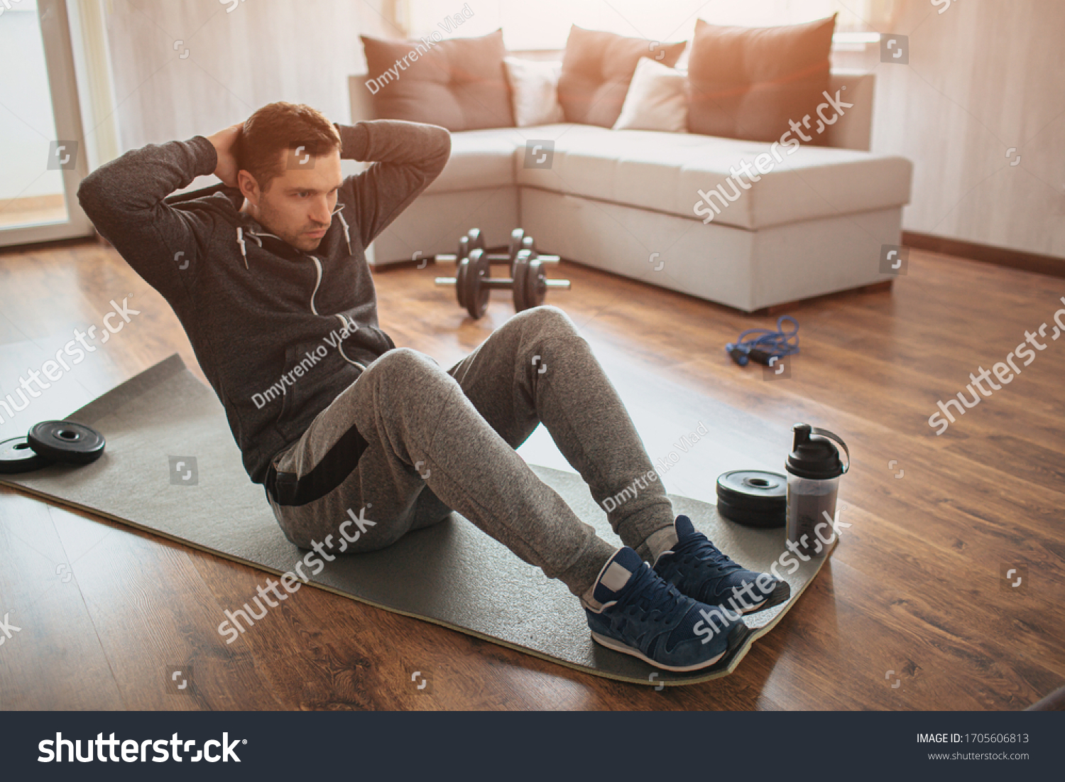 Young ordinary man go in for sport at home. Hardworking freshman sit on mat and do abs excercise. Not easy to start workout alone in apatment. Beginner in action. Sport equipment on floor. #1705606813