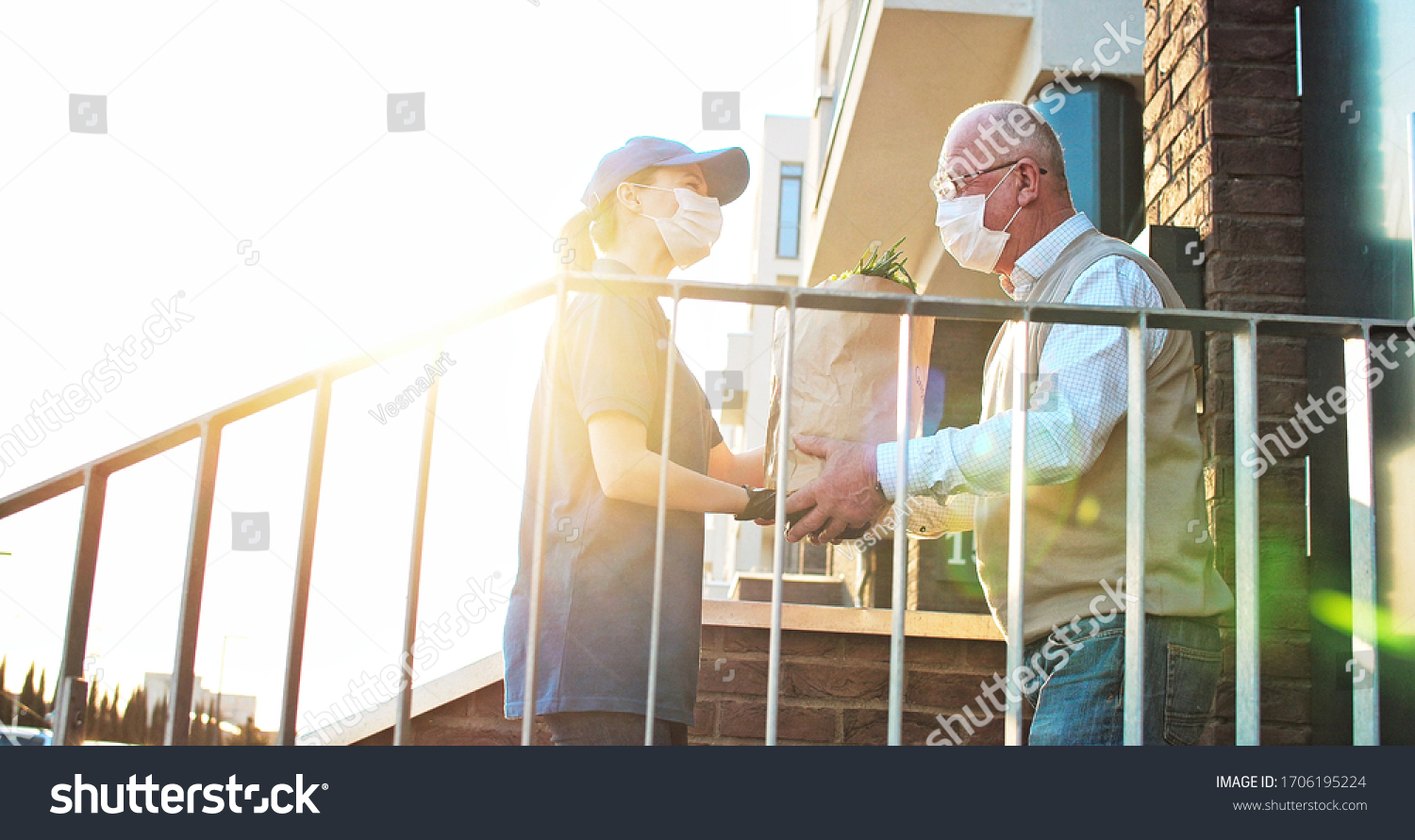 Female courier in medical mask and gloves bringing packet with fresh food to retired old man. Young Caucasian woman handing bag with grocery to male pensioner outdoor at house. Delivery service #1706195224