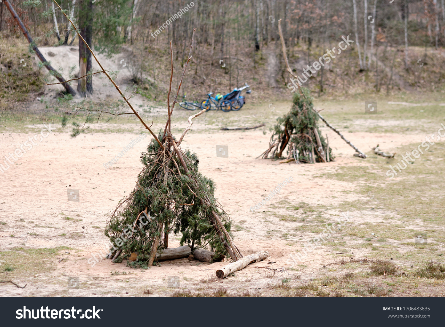 Two tepees built with twigs and branches of conifers standing on sandy ground in the forest with bicycles in the background Seen in Germany in April. #1706483635