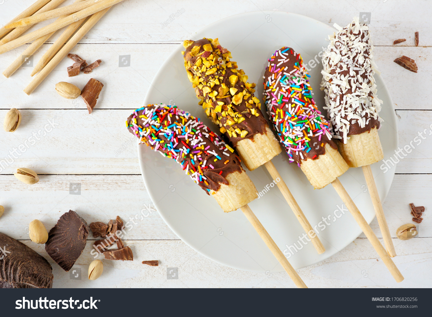 Frozen chocolate dipped banana pops with sprinkles, nuts and coconut. Overhead view table scene on a wood background. #1706820256