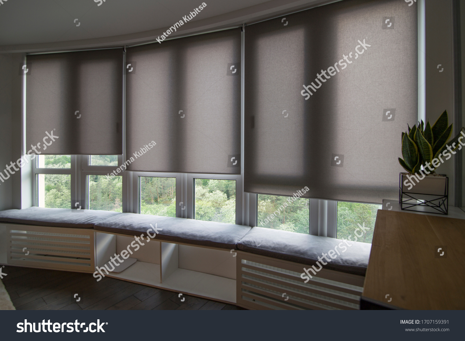Motorized roller shades in the interior. Automatic roller blinds beige color on big glass windows. Home luxury curtaines are above the windosill with pillows. Summer. Green trees outside. #1707159391