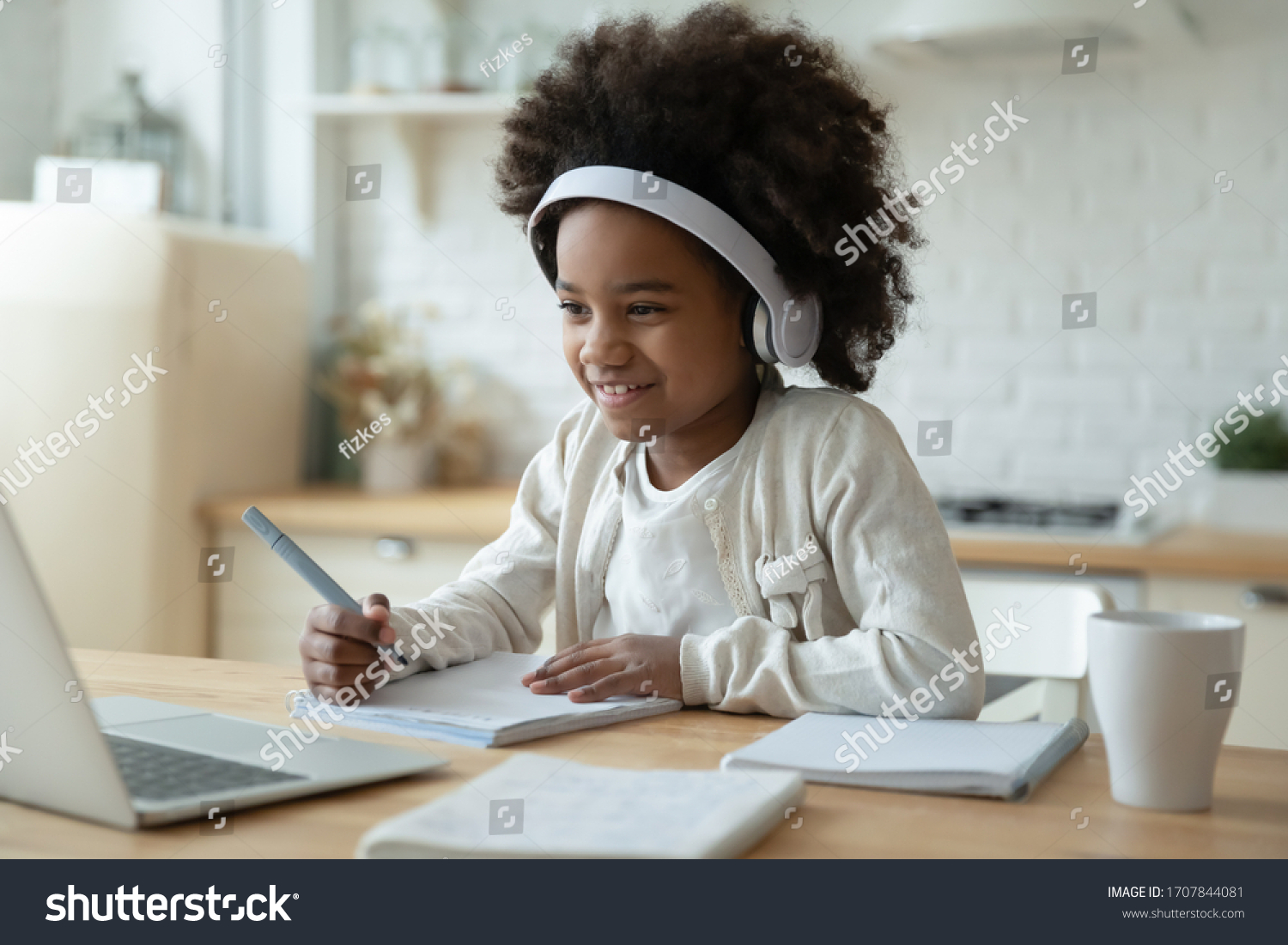 Smiling small African American girl in headphones watch video lesson on computer in kitchen, happy little biracial child in earphones have online web class using laptop at home, homeschooling concept #1707844081