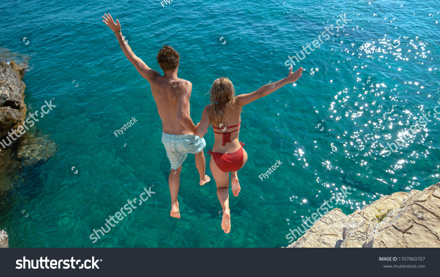 Joyful tourist couple decides to jump off a rocky cliff and dive into sea. Unrecognizable man holds his gorgeous girlfriend's hand while diving into the refreshing blue sea on a sunny summer day. #1707860707