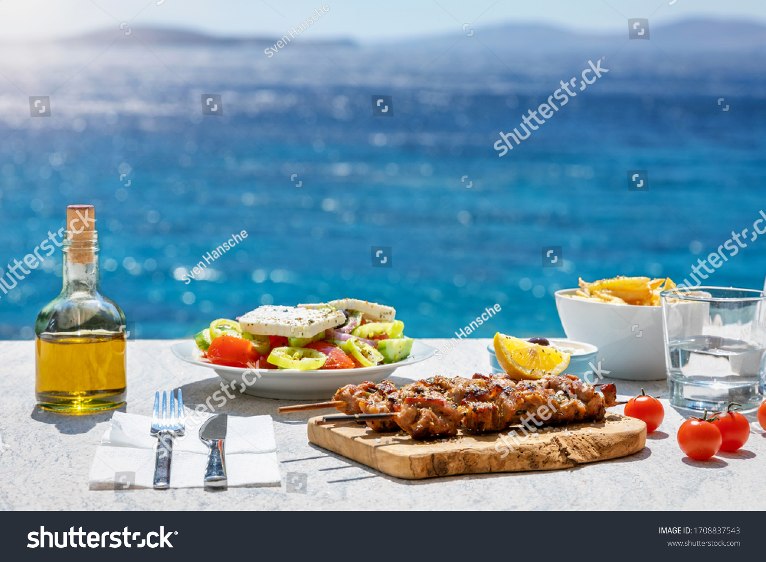 Greek food concept with farmers salad and souvlaki skewers in front of the sparkling, blue Aegean sea during summer time #1708837543