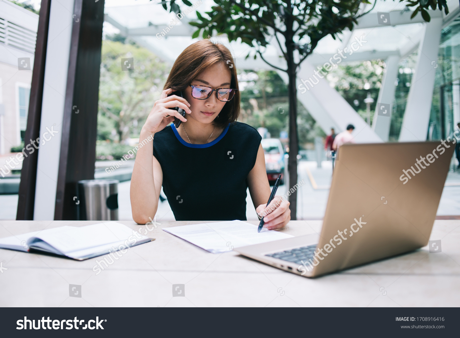 freelance Asian female in black formal wear and eyeglasses sitting at table with notepad holding pen having phone call while using laptop working remotely in workplace on background of glass wall  #1708916416