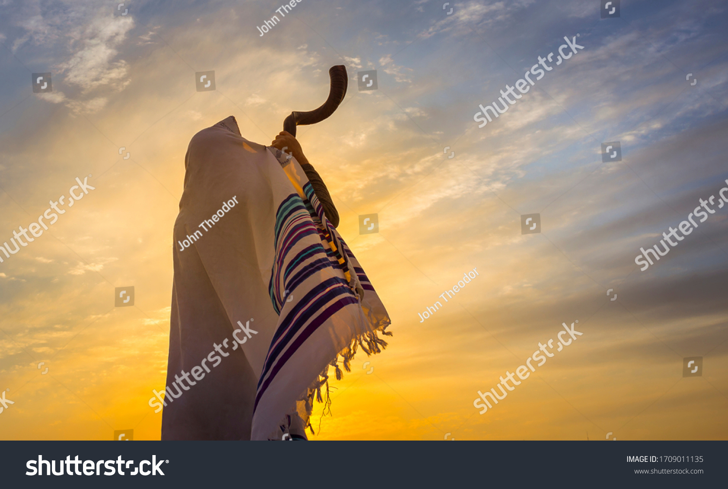 Blowing the shofar for the Feast of Trumpets - Jewish man in a traditional tallit prayer shawl blowing the ram's horn against dramatic sunset sky #1709011135