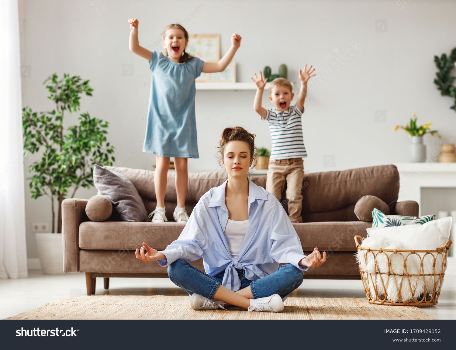 Happy mother with closed eyes meditating in lotus pose on floor trying to save inner harmony while excited children jumping on sofa and screaming in light spacious living room
 #1709429152