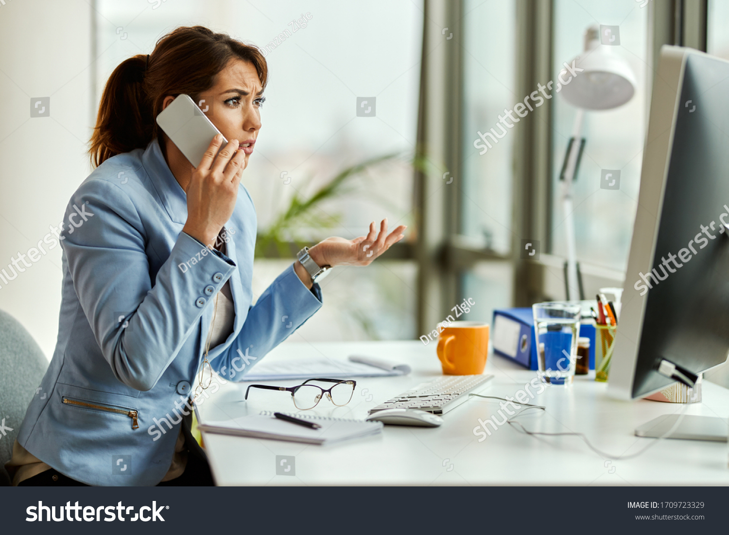 Young displeased businesswoman communicating over mobile phone while working on a computer in the office.  #1709723329