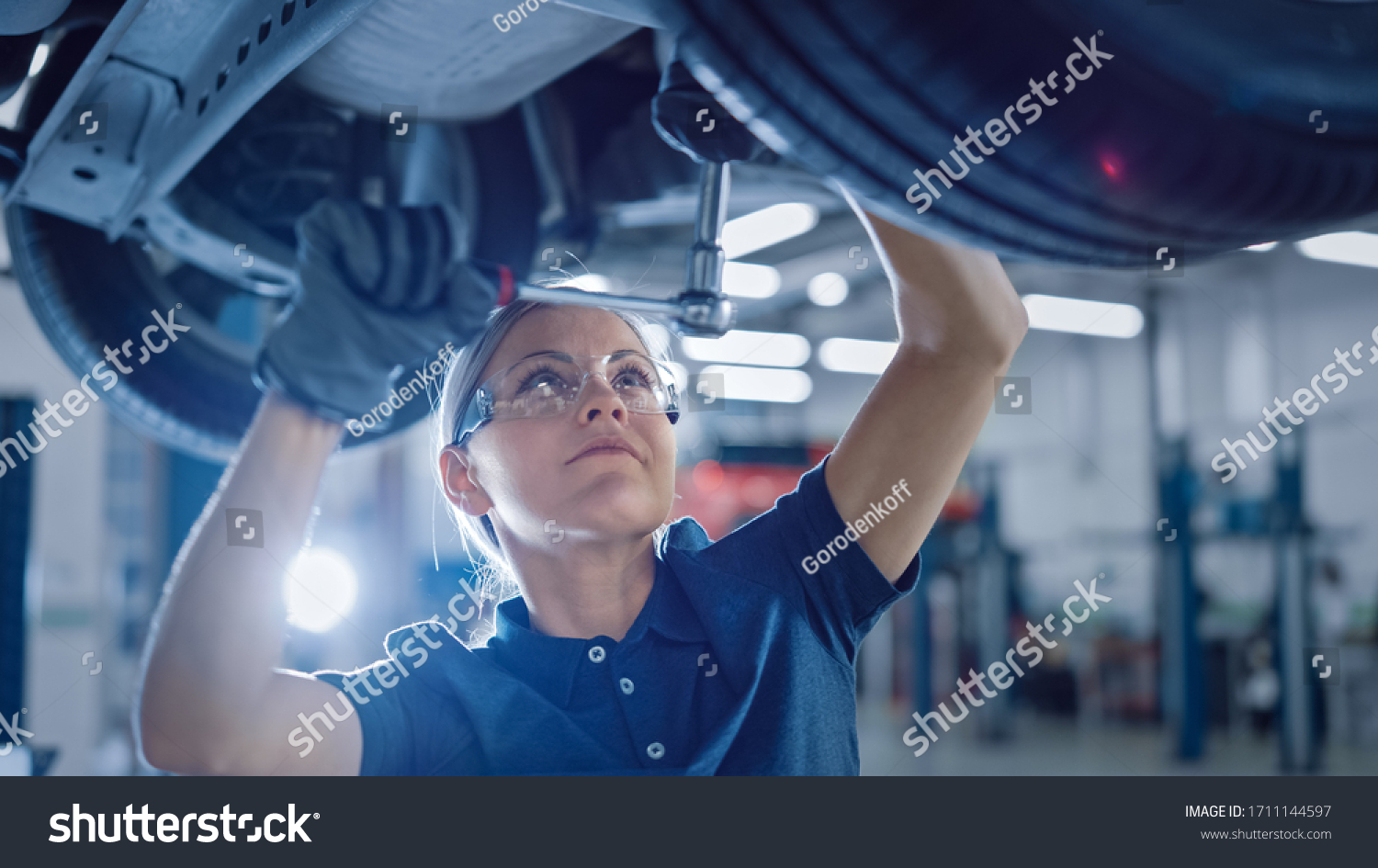 Portrait Shot of a Female Mechanic Working Under Vehicle in a Car Service. Empowering Woman Wearing Gloves and Using a Ratchet Underneath the Car. Modern Clean Workshop. #1711144597