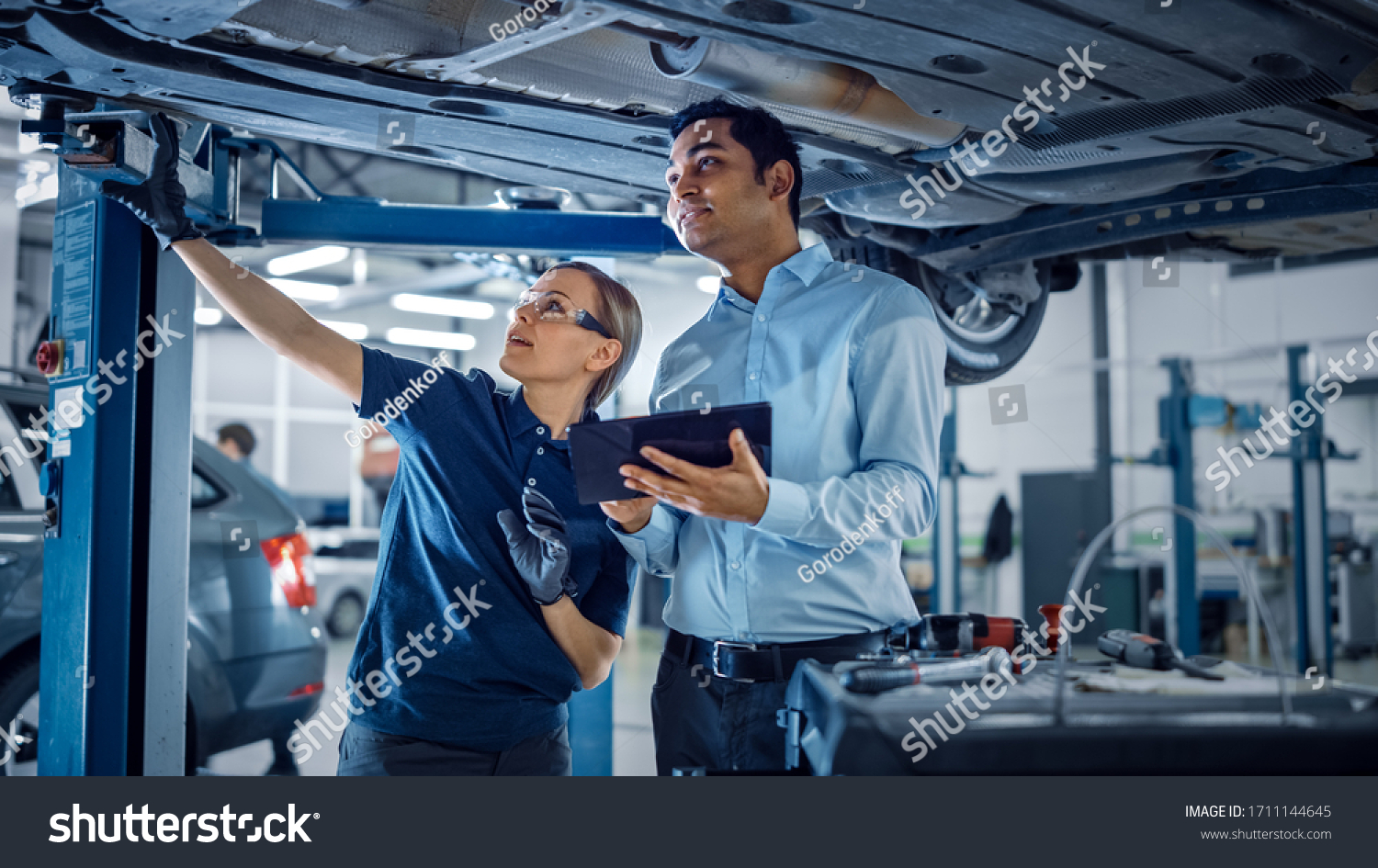 Female Mechanic Talking to a Manager Under a Vehicle in a Car Service. Specialist is Showing Info on a Tablet Computer. Empowering Woman Wearing Gloves and Safety Gloves. Modern Clean Workshop. #1711144645